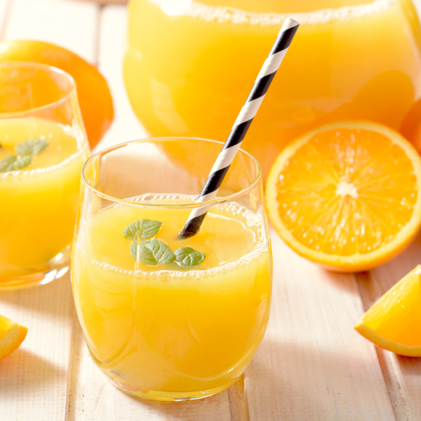 Bicchiere di aranciata con cannuccia bianca a strisce nere e accanto sul tavolo spicchi di arancia e caraffa di aranciata