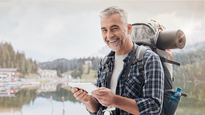 Uomo con zaino sulle spalle davanti a un lago di montagna guarda mappa su tablet e sorride alla fotocamera