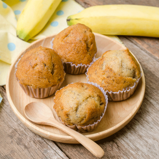 4 muffin alla banana su piatto di legno con cucchiaio di legno e accanto sul tavolo due banane